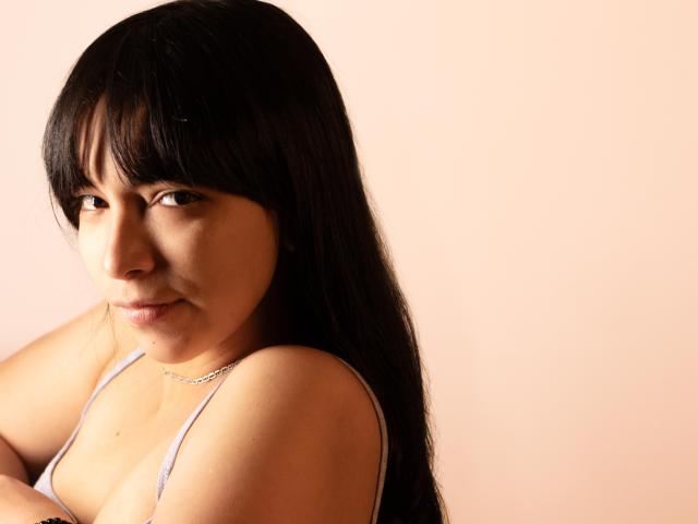 Nessa69s has long black hair, bangs, and a slight smile. She's wearing a white tank top, showing her small tits, and a silver necklace. Her arms are crossed.