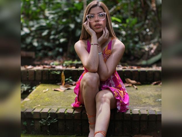 NatyBakers with glasses, pink dress, and orange straps, sitting on mossy brick steps, hands on cheek, looking down, in a forest, showing her tits and pussy in the background.