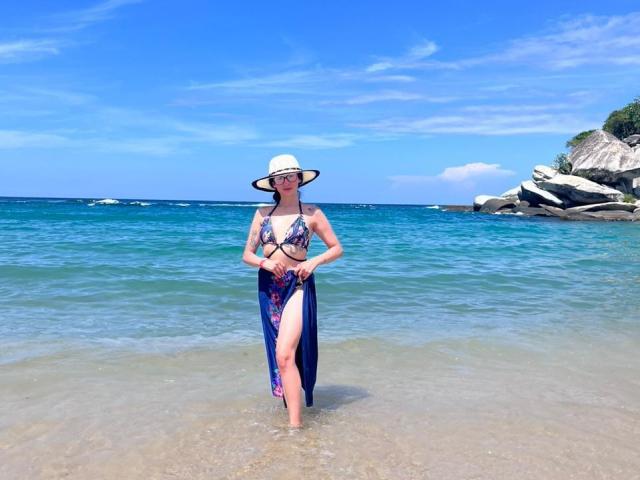 Camille Rios steht im flachen Wasser, trägt einen Blumen-Bikini und einen Sonnenhut, und hat ein durchsichtiges, blaues Kleid an, das bis zu ihrem Oberschenkel reicht und ihre Brüste freilegt. Heller, blauer Himmel, klares Meer..