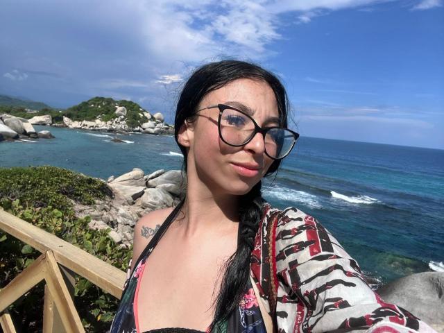 CamilleRios with black braids, glasses, and a patterned shirt, standing by a rocky beach, showing a small tattoo on her shoulder, with a blue ocean in the background.
