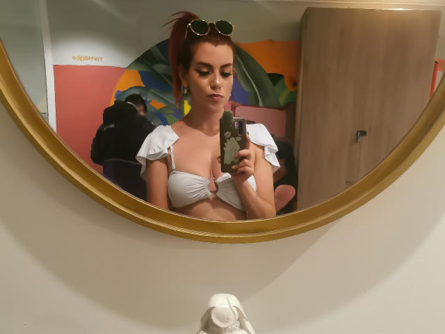 ZoeeMar with red ponytail, wearing white bikini top, holding phone, taking mirror selfie, golden frame, sunglasses on head, subtle makeup, mirror reflection, midriff visible, confident expression.