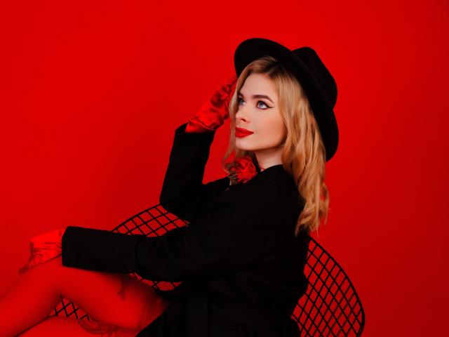 Adele Clifford, con i capelli biondi, un cappello nero, rossetto rosso, un cappotto nero e dei guanti, seduta su una sedia di metallo, che tiene in mano il cappello, su uno sfondo rosso uniforme..