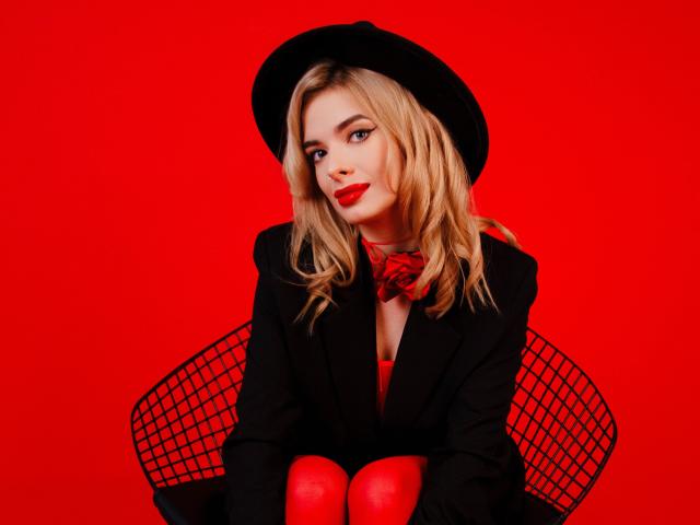 AdeleClifford with blonde hair, red lipstick, black hat, black suit, red bowtie, and red leggings, sitting on a black wire chair against a solid red background.