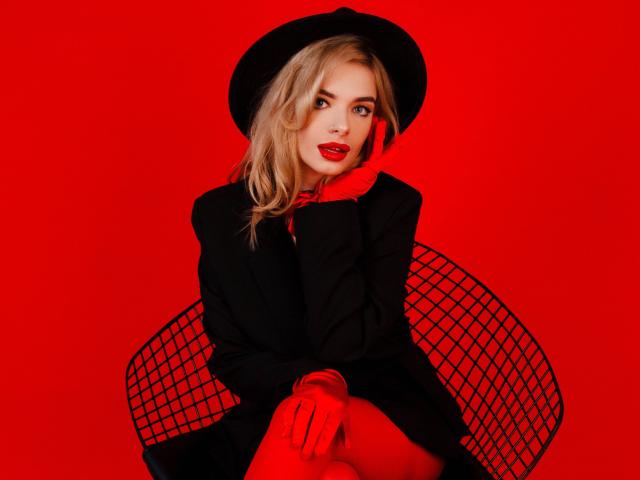 Adele Clifford mit roten Lippen, einem schwarzen Hut, einem schwarzen Anzug und roten Handschuhen, sitzend auf einem Drahtstuhl, blickt in die Kamera, Hände in der Nähe des Gesichts, blondes Haar, roter Hintergrund..