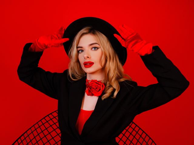 AdeleClifford con cabello rubio, lápiz labial rojo, sombrero negro, guantes rojos, chaqueta negra, vestido rojo, collar de rosas y una silla de malla, sosteniendo el sombrero, fondo rojo, con una expresión segura..