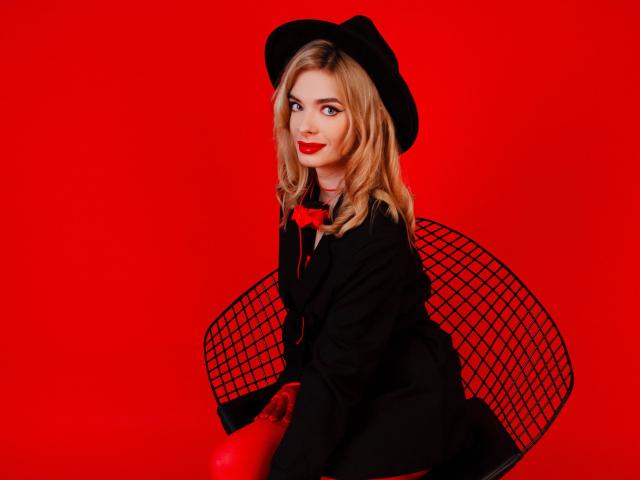 Adele Clifford, com batom vermelho, chapéu preto, terno preto, laço vermelho e luvas vermelhas, sentada em uma cadeira de arame preta, com um fundo vermelho sólido. Suas nádegas estão visíveis..