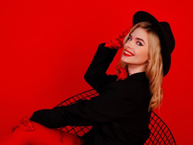 Adele Clifford, com cabelo loiro, batom vermelho, chapéu preto, luvas vermelhas, jaqueta preta, sentada em uma cadeira de arame, sorrindo, fundo vermelho, segurando o chapéu com a mão direita..