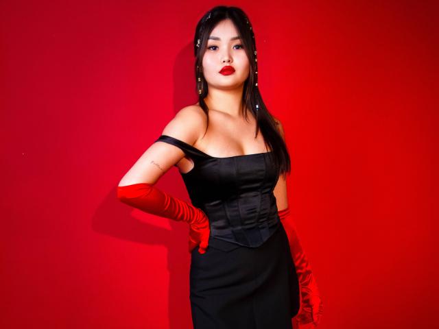 TeyaFaber, avec du rouge à lèvres, une robe noire à une épaule, des gants rouges et des cheveux noirs, debout devant un mur rouge, montrant ses fesses et ses seins dans une pose sexy et confiante..