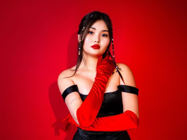 TeyaFaber with red lipstick, black strapless dress, and red velvet gloves, touching her neck, showing off her boobs, against a red background. She's looking at the camera.