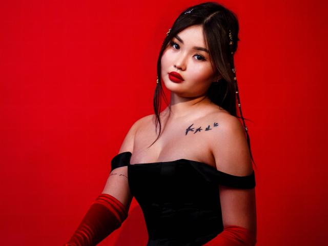 TeyaFaber with red lipstick, black off-shoulder dress, red gloves, and tattoo on left shoulder, looking at camera, red background. Her tits are small, and she has a cute face.