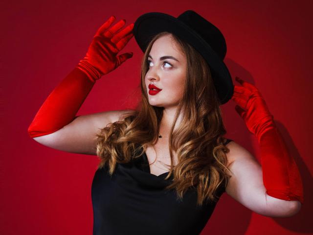 EsterMiln with red gloves, black hat, and red lipstick, looking sideways, has wavy brown hair, black dress, and red background. Her tits are small, and she's sexy.