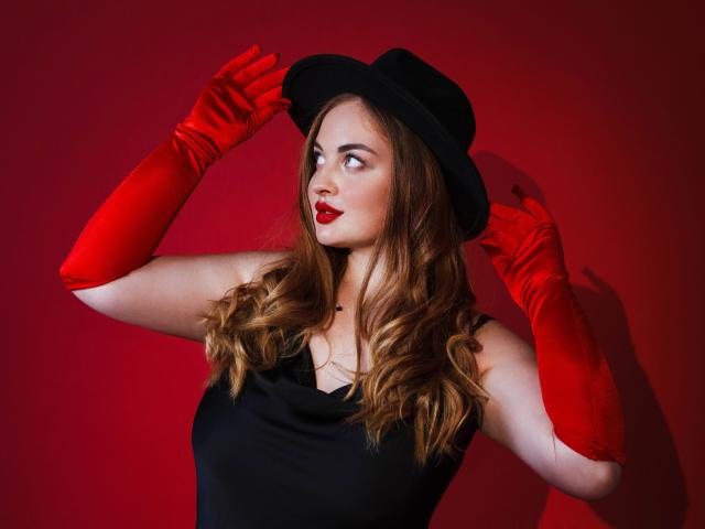 EsterMiln with red gloves, black dress, and hat, holding it with hands on head, has wavy brown hair, red lipstick, and a confident look, against a red background.