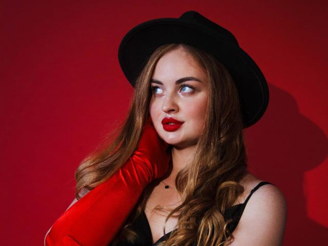EsterMiln with red lipstick, wavy brown hair, black hat, red glove, and black dress, looking sideways with a slight smile. Her tits are small, and she's cute.