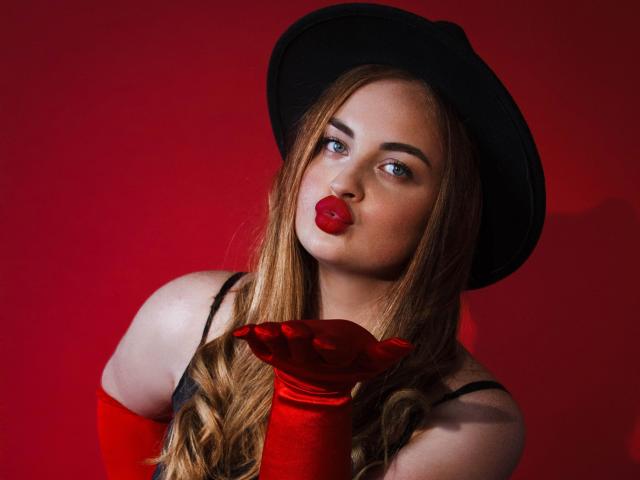 EsterMiln with long blonde hair, black hat, red lipstick, and red satin gloves, pouting with her red lips. Red background. Her tits are partially visible in a black top.