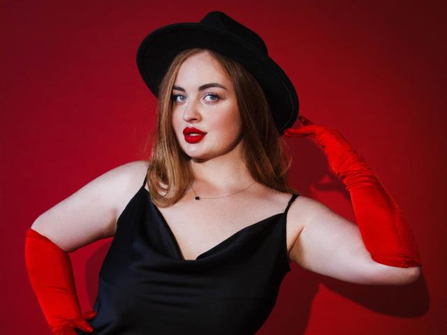 EsterMiln with red gloves, black dress, and hat, holding it, red lips, and a slight smirk, fair skin, and medium tits, against a red background.