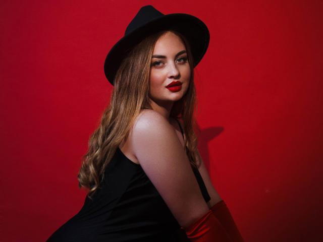 EsterMiln with red lipstick, wearing a black hat and sleeveless black top, showing her tits, and red gloves, against a solid red background, looking over her shoulder.