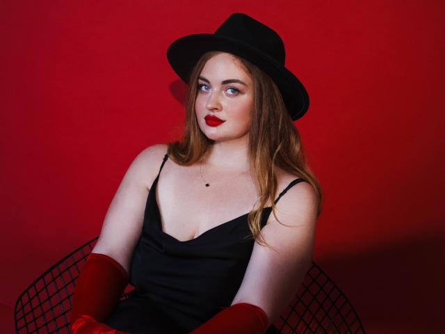 EsterMiln with red lipstick, black hat, and dress, wearing red gloves, sitting in black chair, looking sideways, red background, fair skin, brown hair, small tits, confident.