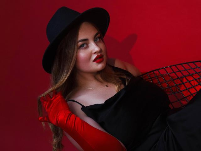 EsterMiln with red lips, black hat, and red gloves, wearing a black dress, sitting against a red wall, holding a black wire basket, showing her boobs.