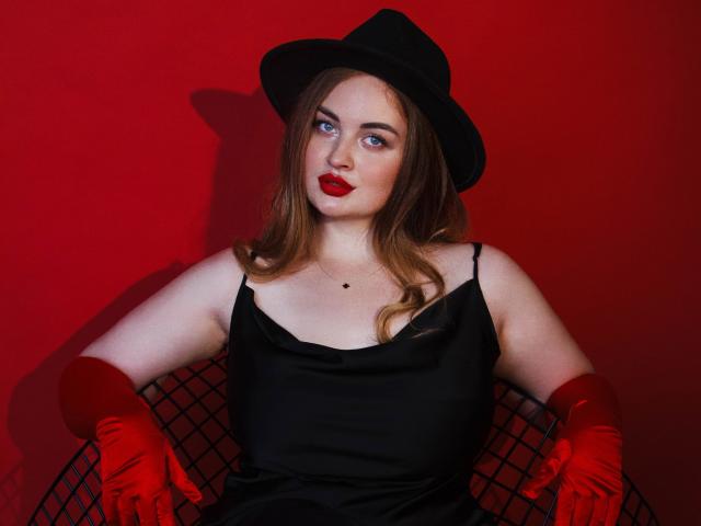 EsterMiln with red lipstick, black hat, black dress, red gloves, and blue eyes, sitting with arms on a black wire chair, showing her tits and a small black mole.