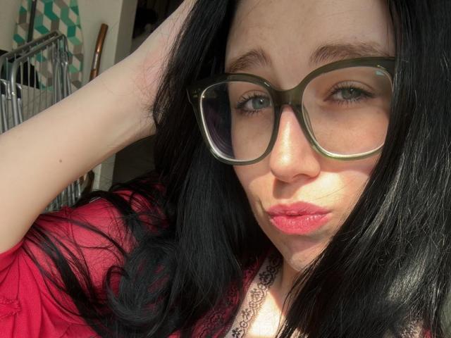 Macdi has black hair, green glasses, and pink lipstick, pouting with her hand in her hair, wearing a red shirt and lace collar, sunlight on her face.