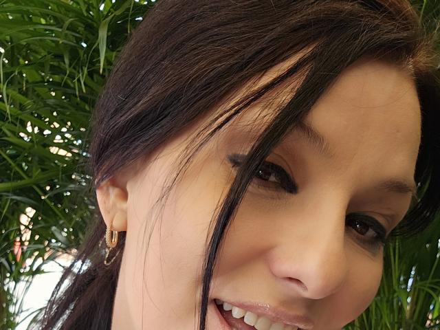 JoliDanielle's face with dark hair, brown eyes, small gold hoop earring, and a slight smile showing her teeth. Green foliage in the background. She's looking to the right.