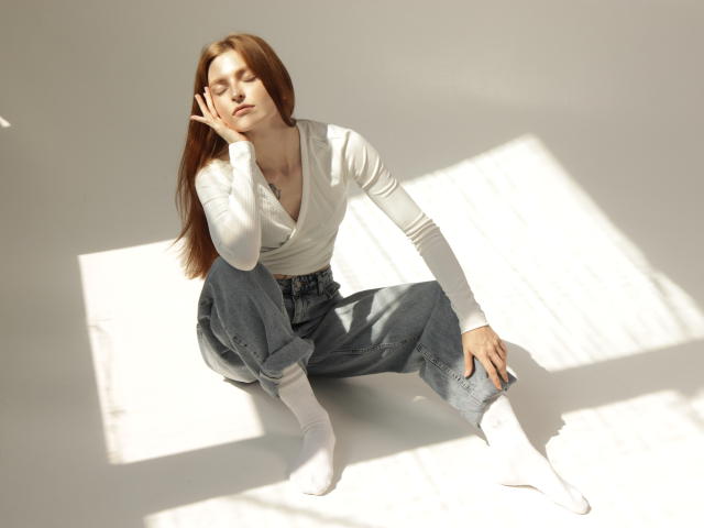 Malena Golden, com cabelos ruivos longos, uma blusa branca e jeans azuis, está sentada no chão, com os olhos fechados, a mão no rosto, meias brancas, e a luz do sol projetando uma sombra. Seios visíveis sob a blusa..