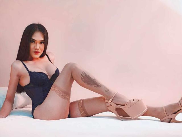 SexyTemptacious with long black hair, wearing a black lace bodysuit, beige thigh-high stockings, and pink platform heels, showing a tattoo on her right thigh, sitting on a bed.