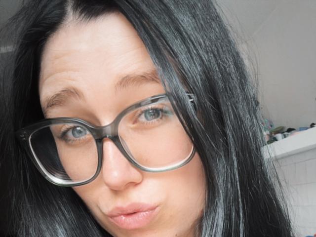 Macdi has black hair, blue eyes, and wears big, black, rectangular glasses. She's pouting with slightly parted pink lips. Her nose is small and she's fair-skinned.