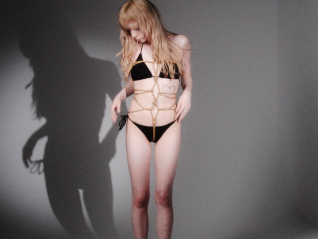 LisaMetalHoney with long blonde hair, black bikini, and gold rope body harness, looking down, shadow on gray wall, standing, slim, small tits, black panties, pale skin.