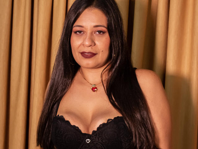 MiaCallie with long dark hair, wearing a black lace bra, showing her tits, medium brown skin, dark red lipstick, gold necklace with red pendant, smiling slightly.