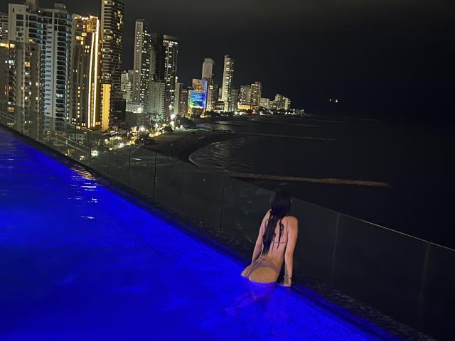 SofiMonroy with long dark hair, topless, wearing yellow panties, sitting on edge of blue-lit infinity pool, facing city skyline at night, black glass fence to her right.