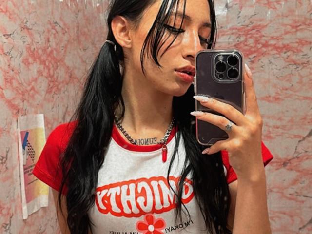 SofiMonroy with black pigtails, red and white rag tee, holding phone, silver necklace, and long nails, pouting slightly, taking a selfie in a pink marble bathroom.