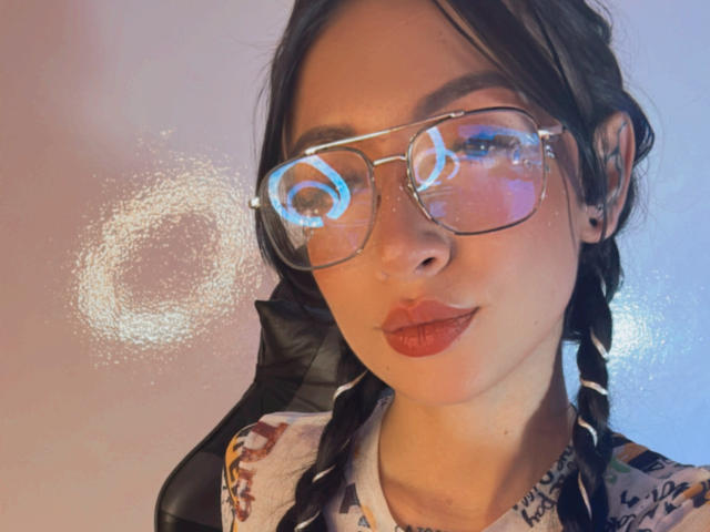 SofiMonroy with glasses, reflecting a light circle on her face, has braided hair, wearing a floral shirt, and red lipstick, sitting against a white wall.