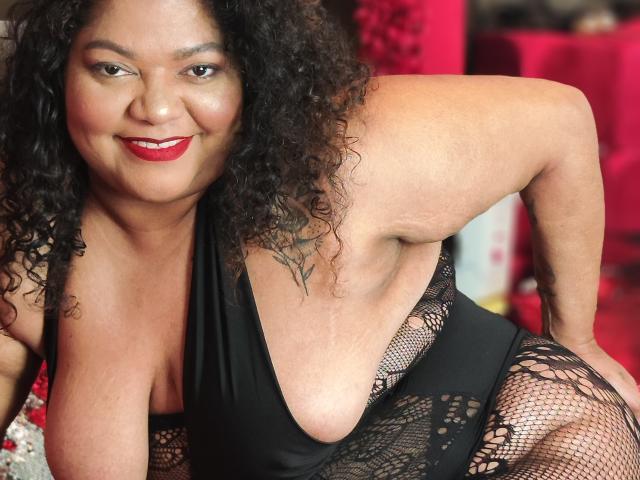 BrunetteBlue with curly black hair, red lipstick, smiling, wearing a black lace lingerie top, exposing her large tits and showing a tattoo on her right arm.
