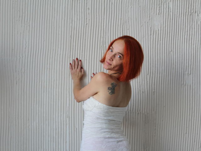BreeTanya with red hair, white strapless dress, tattoo on right shoulder, red nails, facing wall, looking back, smiling, hands on wall, white textured background.