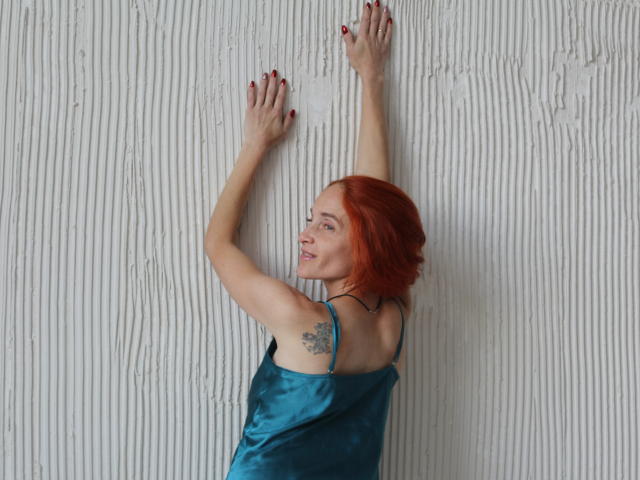 BreeTanya with red hair, blue satin dress, arms raised, hands against white textured wall, tattoo on right shoulder, red nails, looking to the side, slight smile.