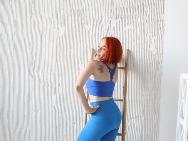 BreeTanya with red bob has blue sports bra and high-waisted blue leggings, hands on hip, tattoo on right shoulder, wooden ladder behind her, smiling, light skin.