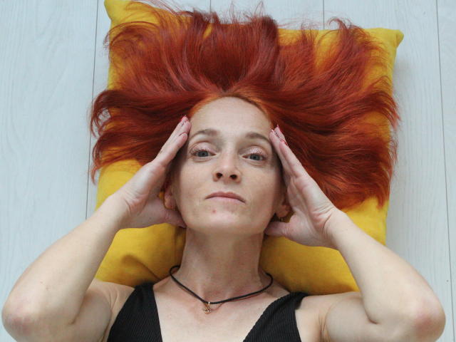 BreeTanya with red, fanned-out hair, black top, yellow pillow, hands on her head, wearing a black necklace, looking slightly bored, pale skin, red nails, subtle freckles.