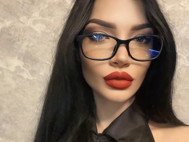 MoiraMilicent, con i suoi lunghi capelli neri, indossa occhiali neri e rossetto rosso, ha un viso carino ed è rivolta direttamente verso la fotocamera con un leggero sorriso..