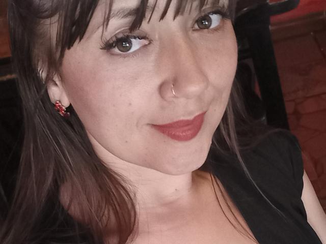 LaFrancaiseJessy with wavy brown hair, red lipstick, and a blue dress exposing her tits and small brown nipples. She's leaning forward, looking at the camera with a slight smile.