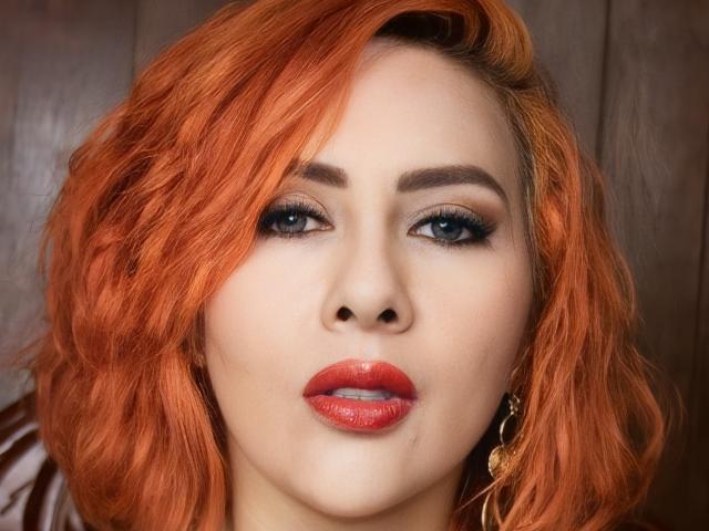 El rostro de NikiiFoxy, con cabello rojo brillante y ondulado, ojos azules, cejas oscuras, lápiz labial rojo y aretes dorados. Su maquillaje es ahumado, y tiene una ligera y sensual sonrisa..