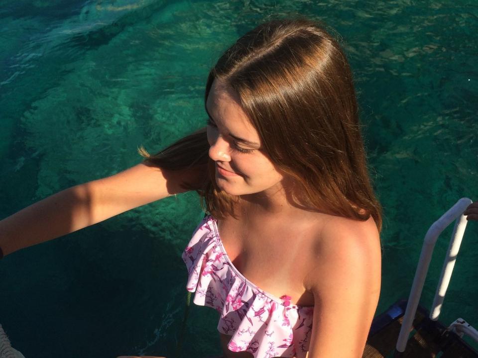 LivesEro with long brown hair, small tits, and a pink floral strapless top, standing by a green pool, looking down with a slight smile, sunlight on her face.