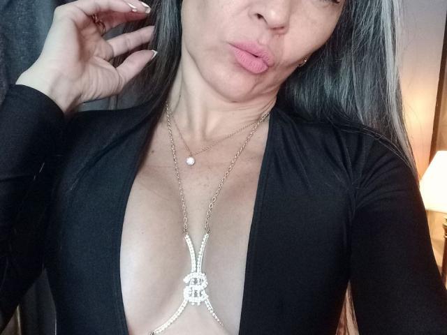 Nicolette Shea, com lábios carnudos, vestindo uma blusa preta com decote profundo, mostrando seus seios, um colar de prata, cabelos longos e grisalhos, e um anel no dedo..