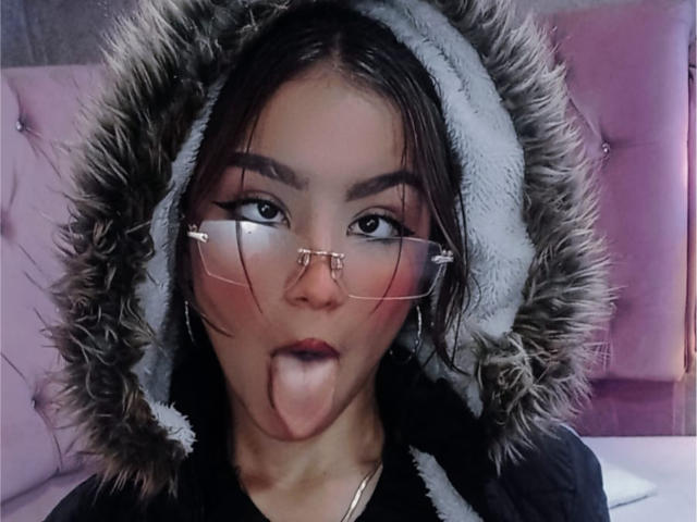 Ashley Miller mit Brille, Zunge heraus, trägt eine pelzige, graue Kapuze, blickt nach oben mit einem verspielten Ausdruck, dunkles Haar und einem rosafarbenen Schimmer auf ihren Wangen..