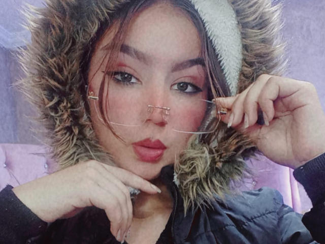 AshleyMiler with a puffy jacket, wearing a fur-lined hood, holding glasses in her right hand, pink eyeshadow, red lipstick, and a cute nose ring, looking at the camera.