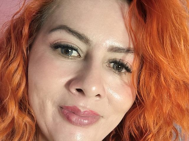 O rosto de NikiiFoxy, com pele brilhante, cabelo encaracolado laranja e lábios rosados e brilhantes, olhando ligeiramente para o lado com um sorriso discreto. Seus olhos verdes têm um leve brilho..