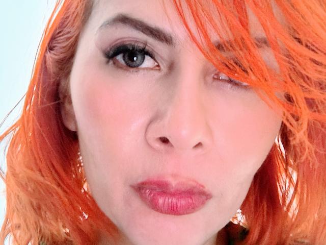 NikiiFoxy has orange hair, one eye winking, and pink lipstick on her slightly parted, shiny lips. Her fair skin and black eyeliner stand out against her bright hair.