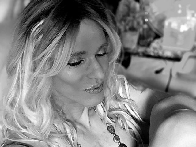 Black and white photo of Cherris with blonde wavy hair, downcast eyes, glossy lips, and a necklace, sitting with her arm raised, showing her tits.