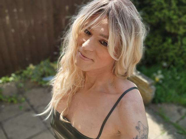 Blonde Cherris with wavy hair, small tits, black spaghetti strap top, tattoo on upper arm, smiling, outdoor sunlight, greenery in background.