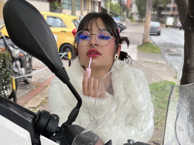 LaraAhB con un caramelo rosa en la boca, vistiendo una chaqueta blanca esponjosa, gafas redondas, auriculares y un piercing en la nariz, sentada en una motocicleta, con una acera y un coche amarillo al fondo..